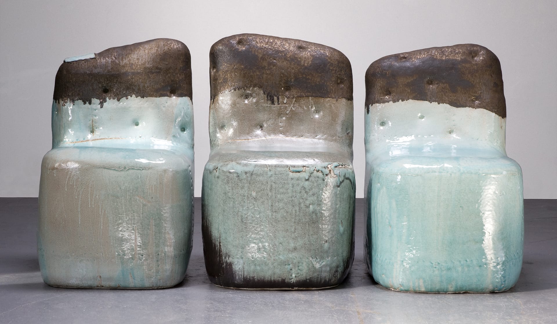 Three abstract, block-shaped ceramic chairs with a gradient of brown, turquoise, and light blue colors on a gray background.
