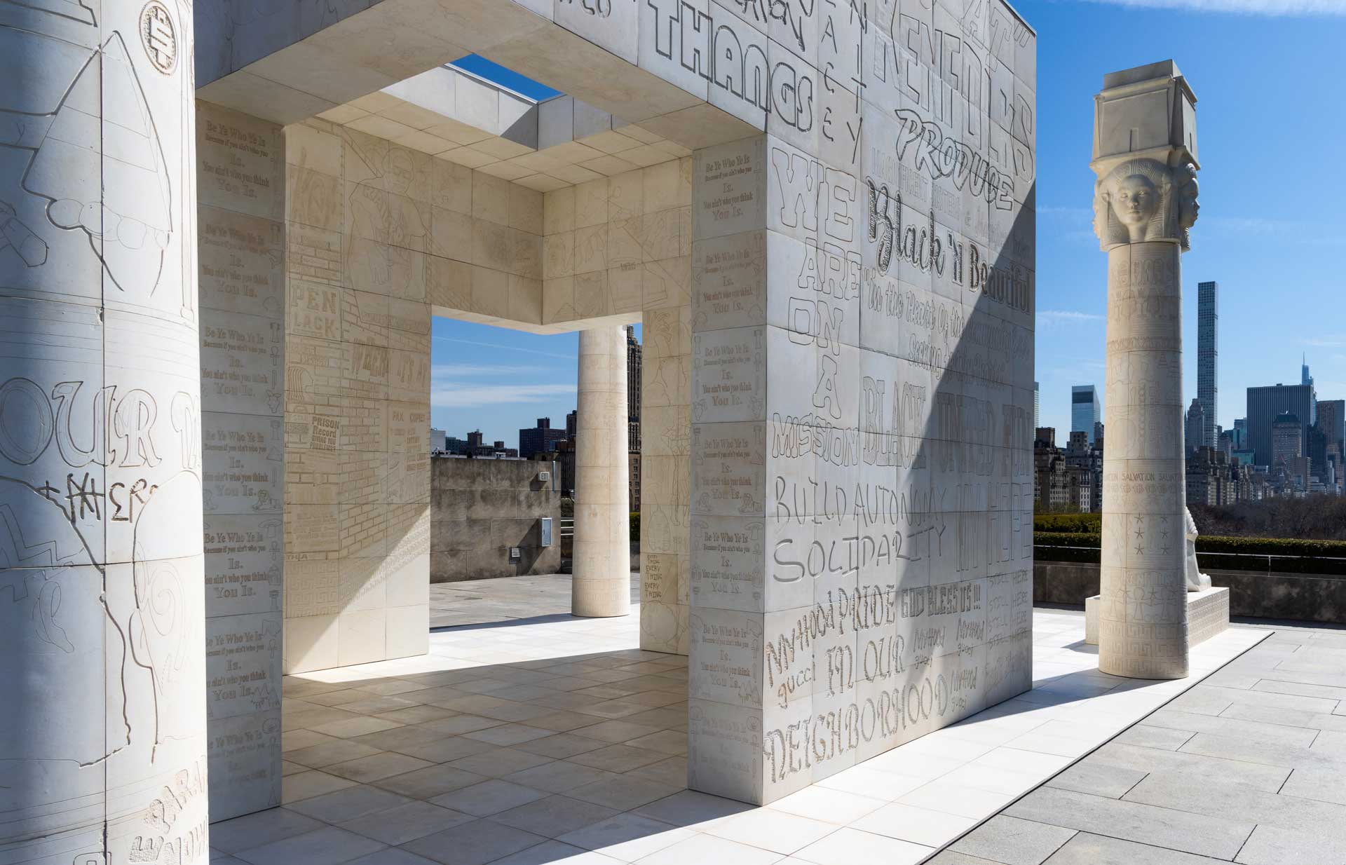 Stone structure with engraved text and columns under a clear blue sky, city skyline visible in the background.