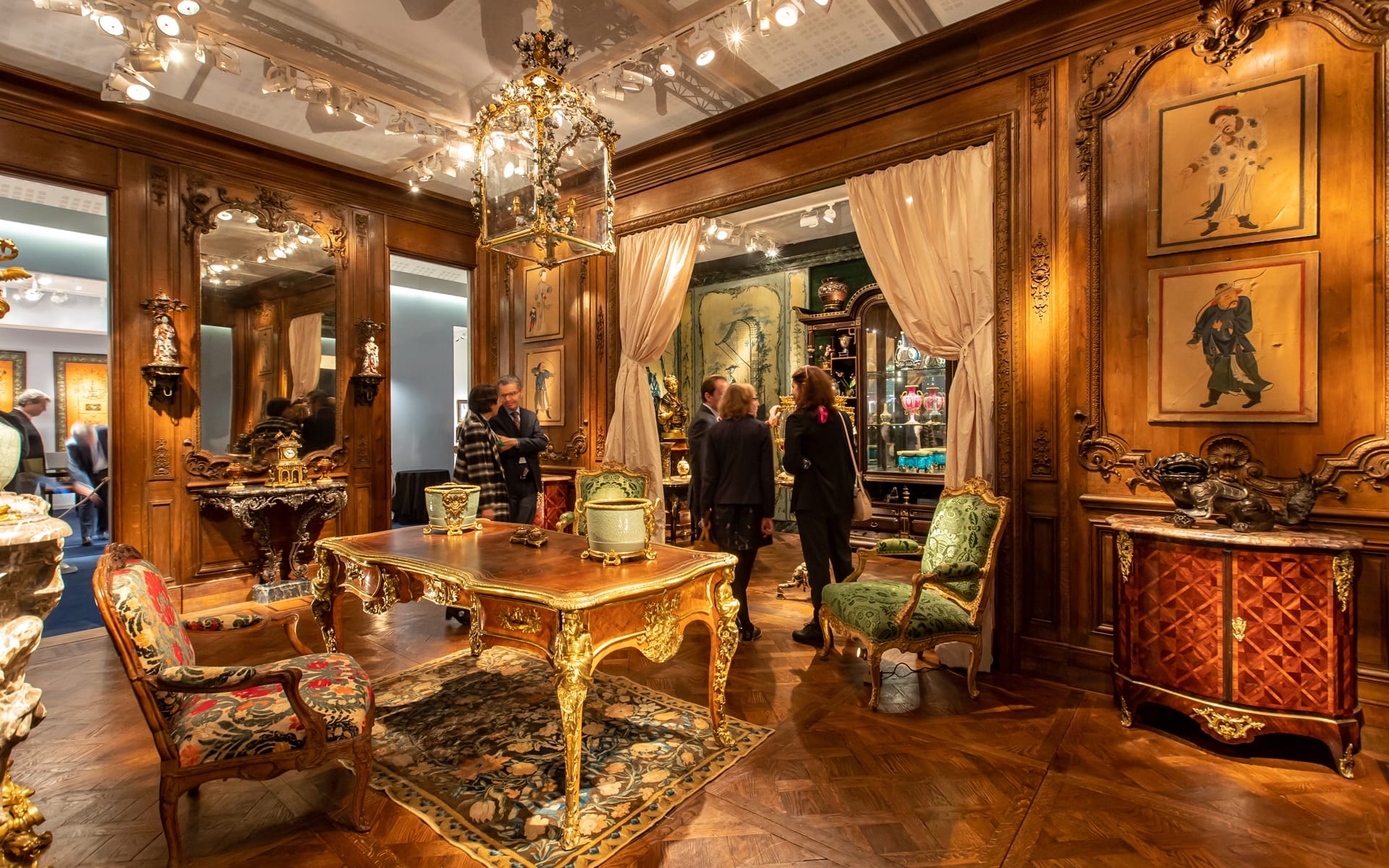 Luxurious antique room with ornate wooden furniture, decorative paintings, and people admiring the elegant decor.