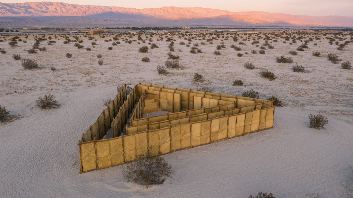 Aerial view of a triangular bamboo maze in a desert landscape with mountains in the background during sunset.