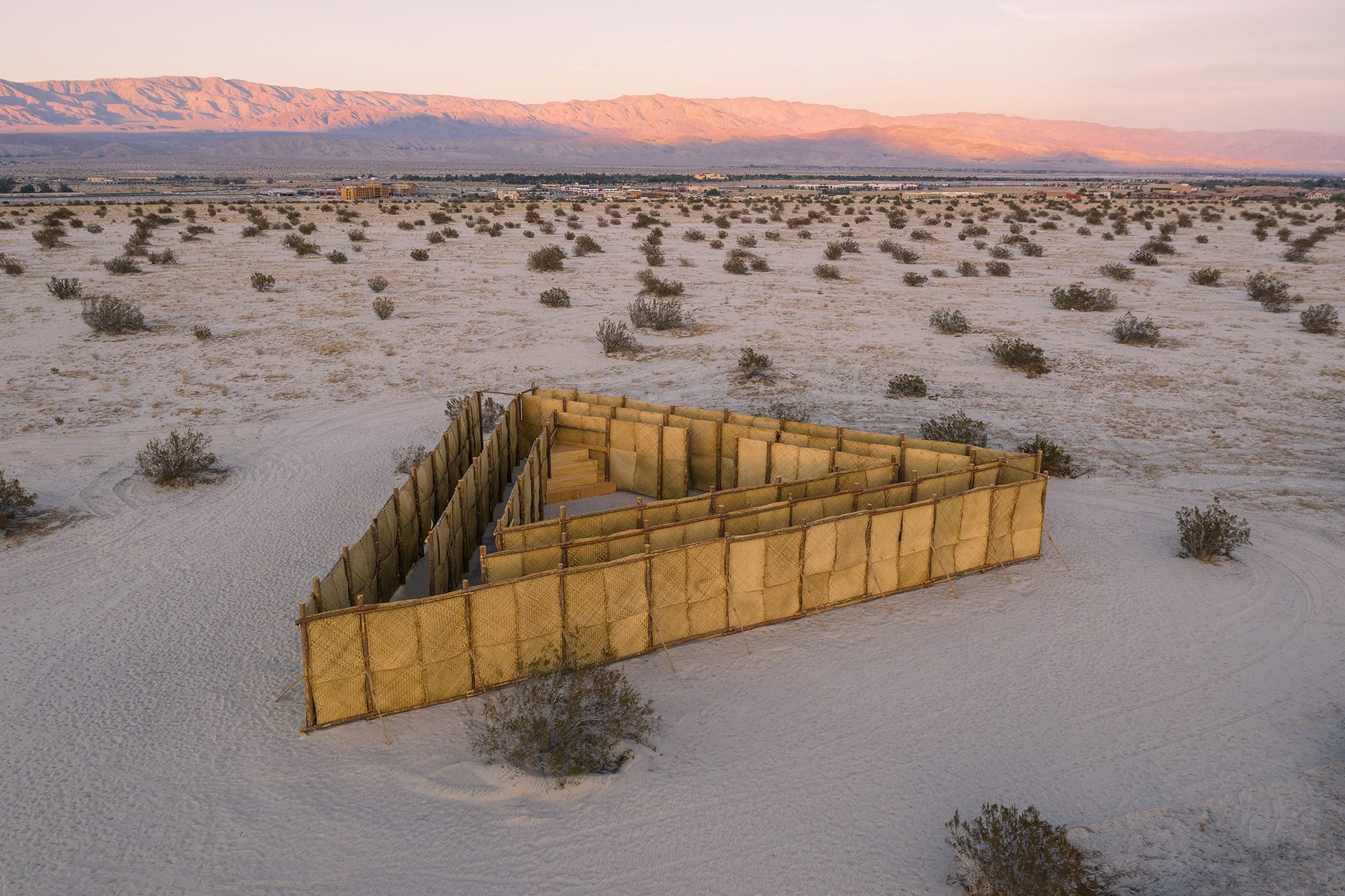Aerial view of a triangular bamboo maze in a desert landscape with mountains in the background during sunset.