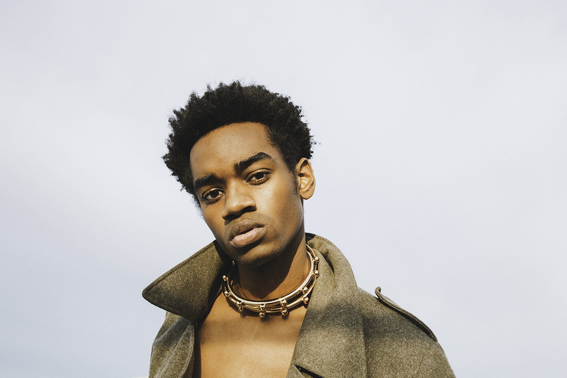 Person wearing a thick necklace and a coat, looking into the camera against a clear sky background.