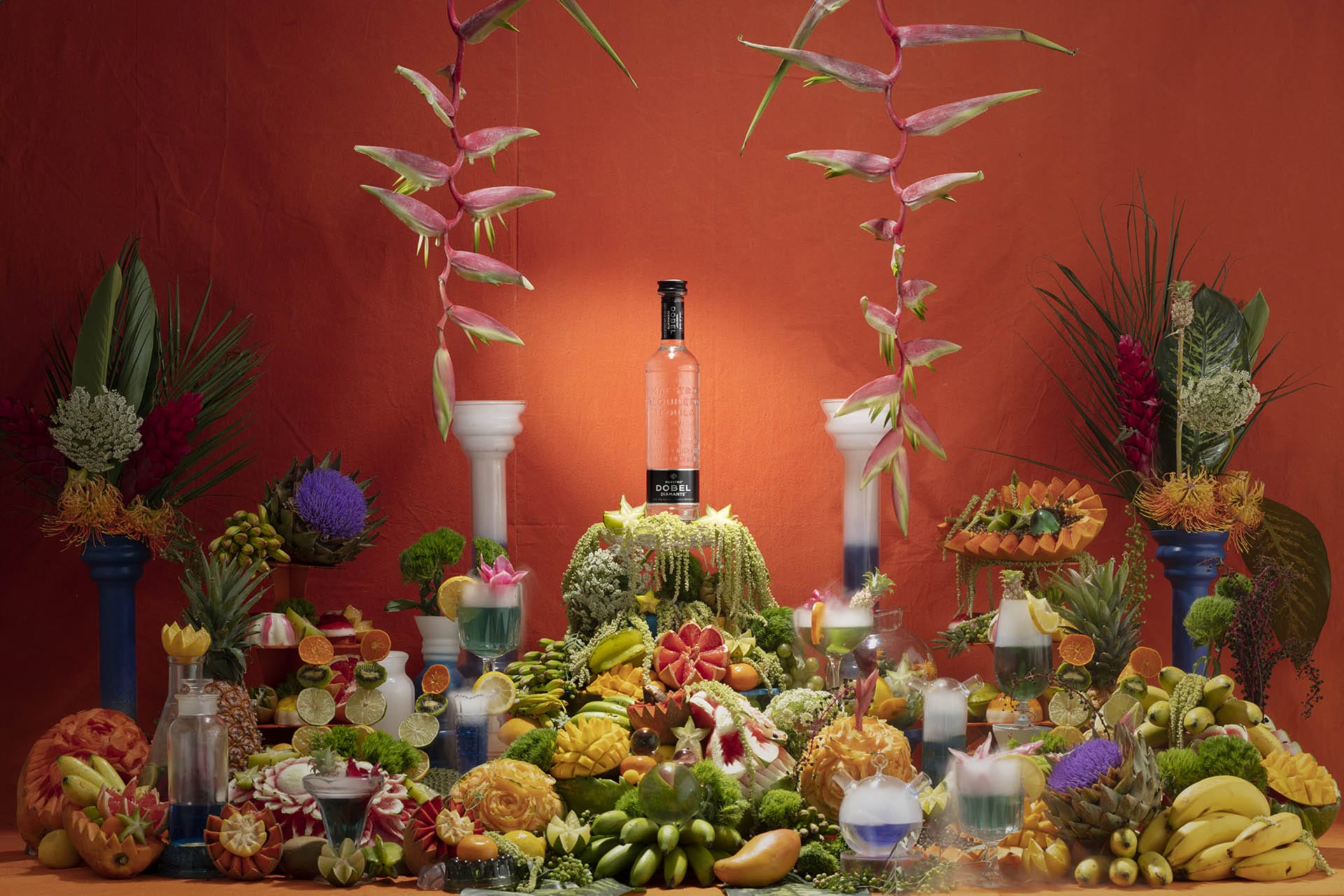 An array of tropical fruits and flowers displayed around a bottle on a vibrant orange backdrop.