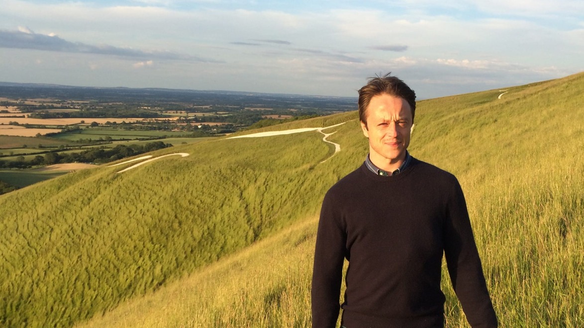 Man standing on a grassy hill with a scenic landscape view in the background under a clear blue sky.