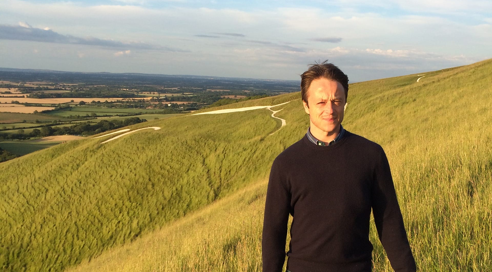 Man standing on a grassy hill with a scenic landscape view in the background under a clear blue sky.