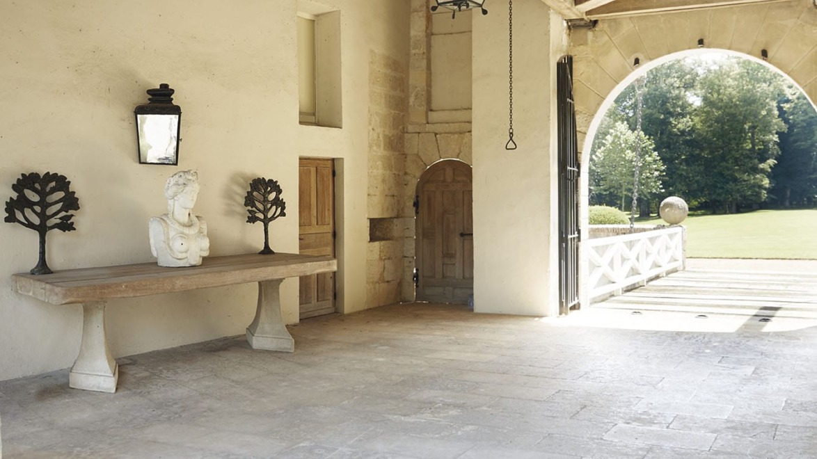 Rustic white stone room with a wooden table, stone bust, metal trees, large lantern, and arched entry to a green garden.