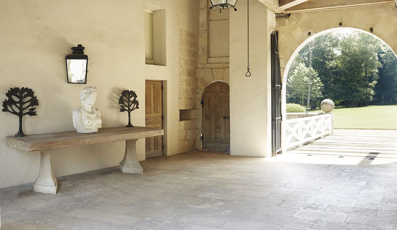 Rustic white stone room with a wooden table, stone bust, metal trees, large lantern, and arched entry to a green garden.