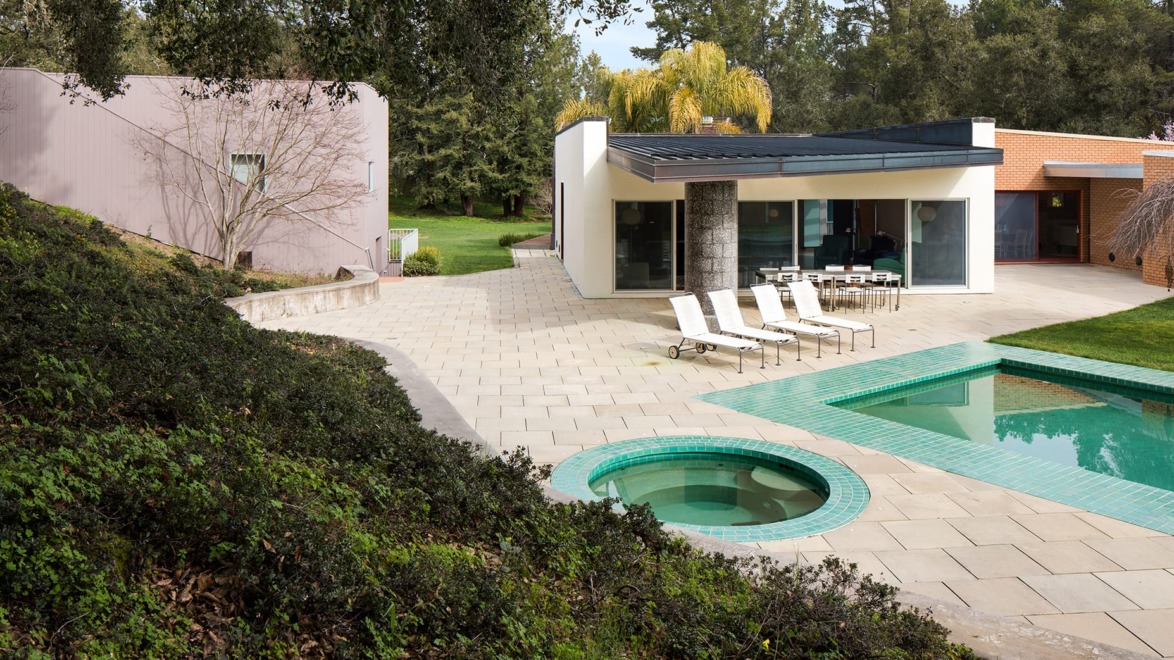 Modern house with a pool and patio, surrounded by greenery and trees on a sunny day.