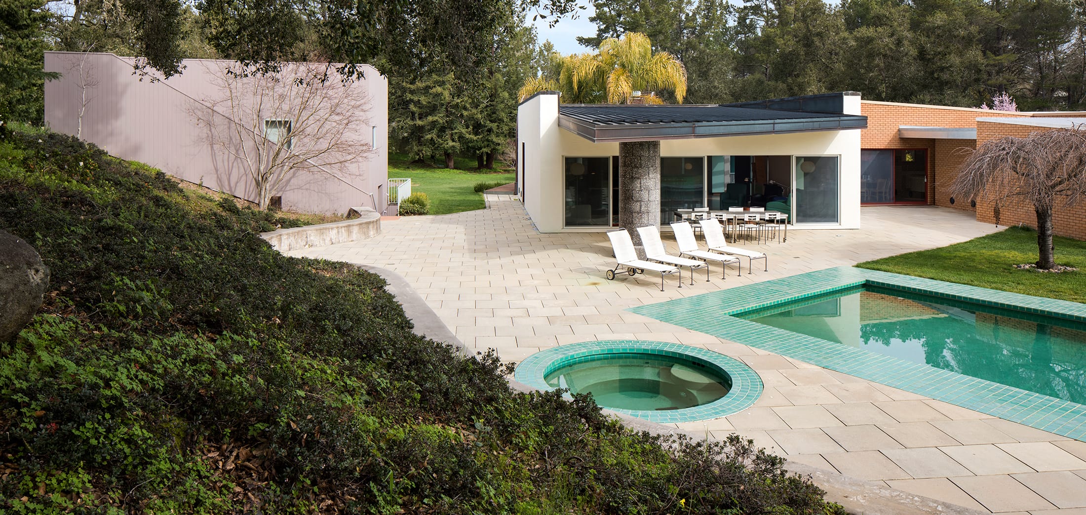 Modern house with a pool and patio, surrounded by greenery and trees on a sunny day.