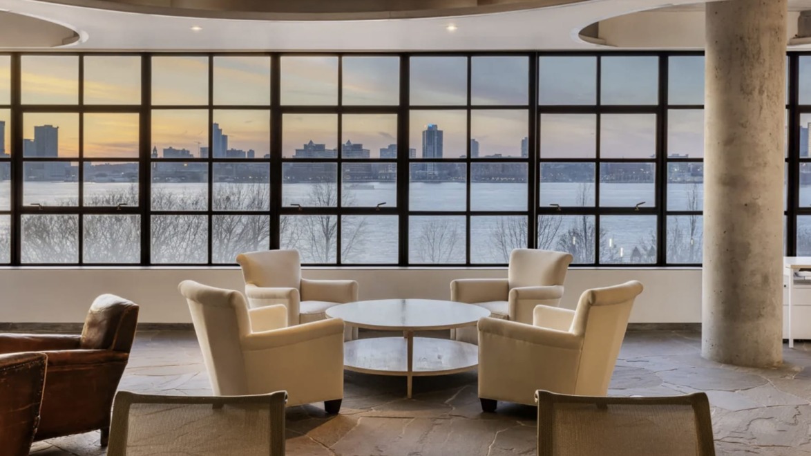 Modern office lounge with four white chairs around a table, large windows showcasing a city skyline and river view at sunset.