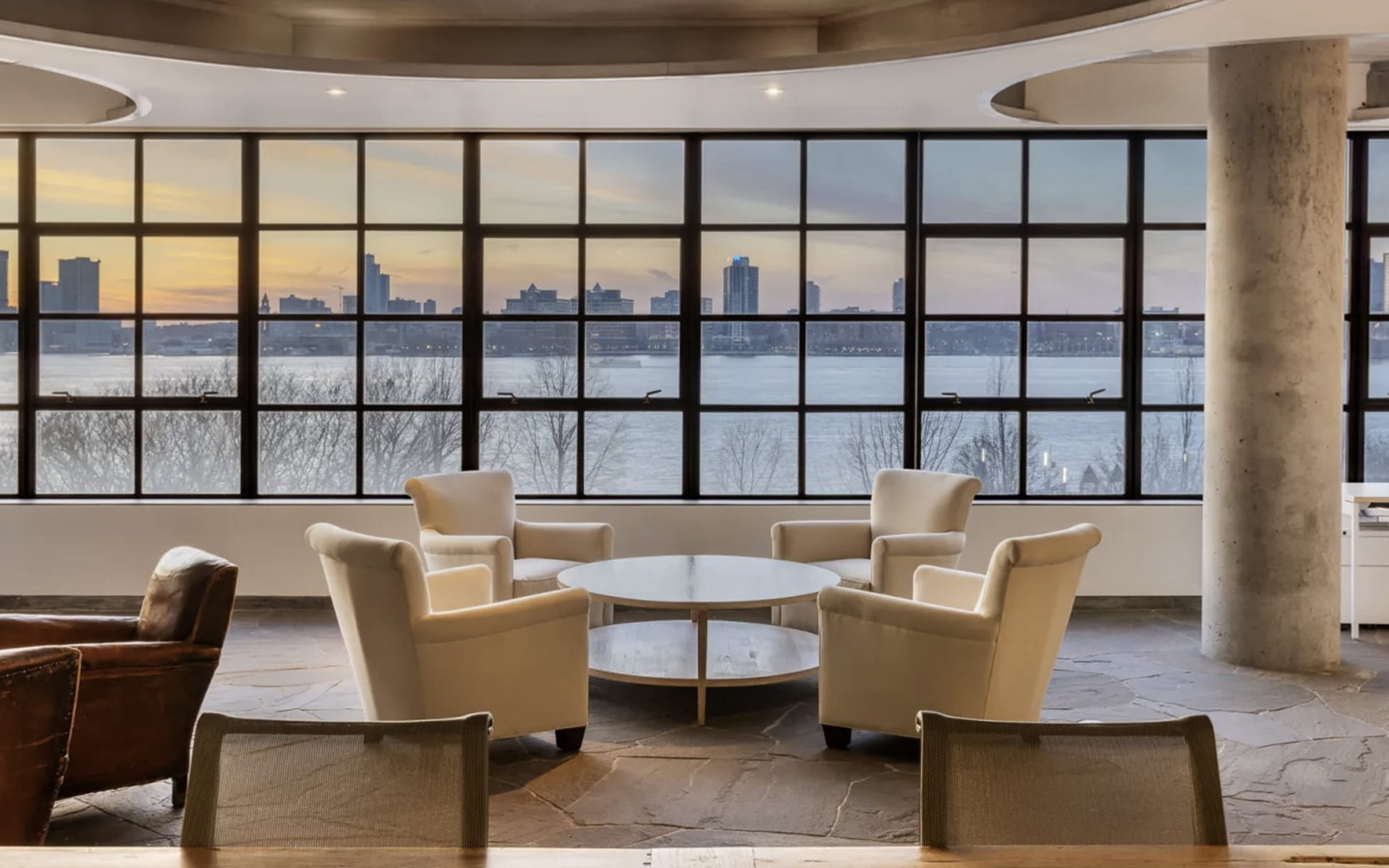 Modern office lounge with four white chairs around a table, large windows showcasing a city skyline and river view at sunset.