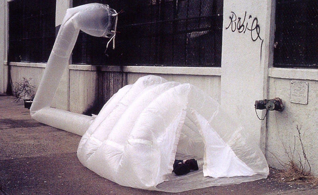 Inflatable igloo-like shelter attached to a wall, with a long air duct and graffiti visible in an urban setting.