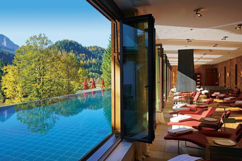 Luxurious spa room with panoramic mountain views and an indoor pool surrounded by lounge chairs in natural sunlight.