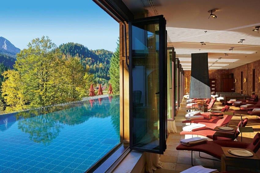 Luxurious spa room with panoramic mountain views and an indoor pool surrounded by lounge chairs in natural sunlight.