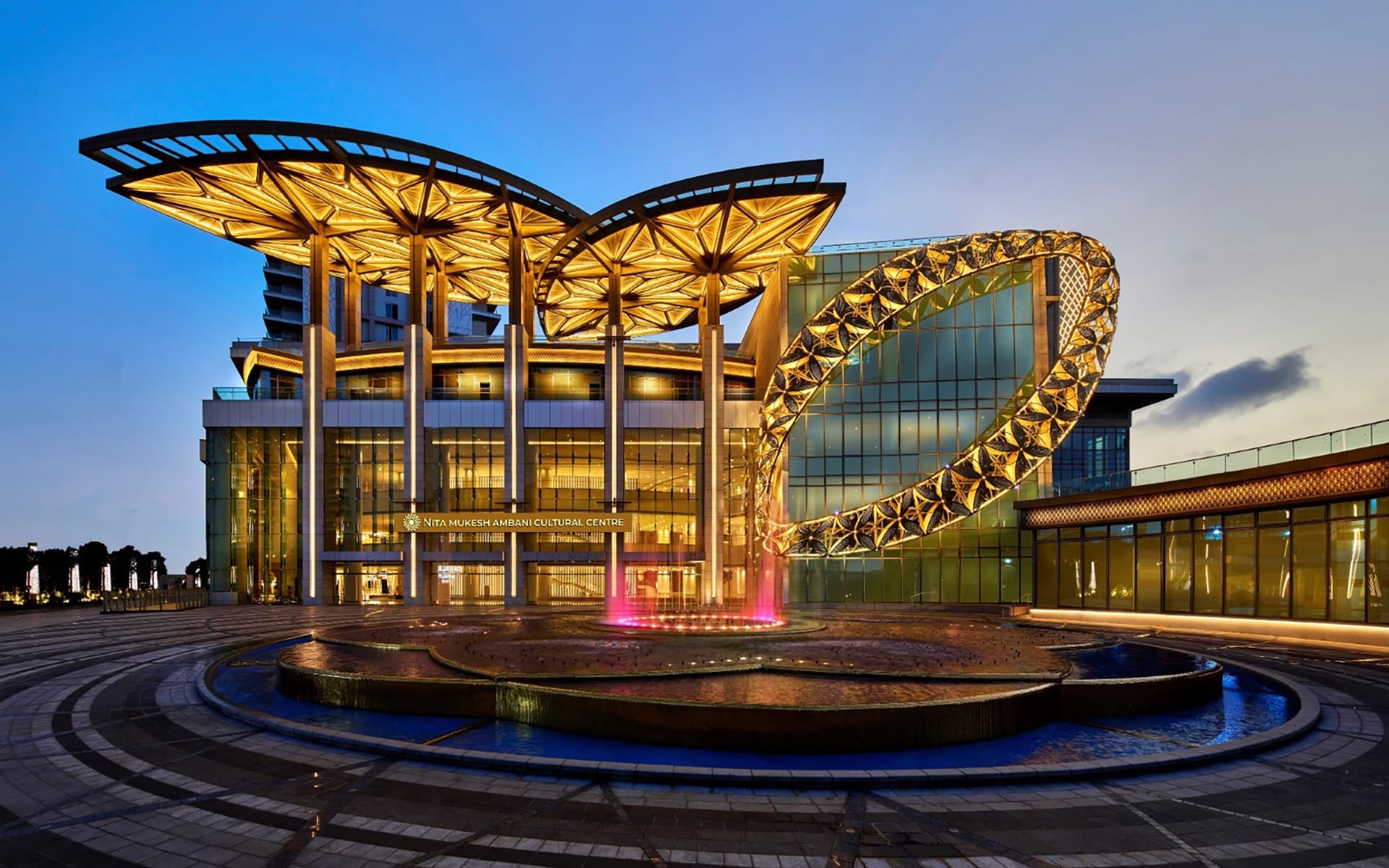 Exterior view of Noor Al Aser Cultural Centre with golden illuminated structure and fountain in the foreground at dusk.