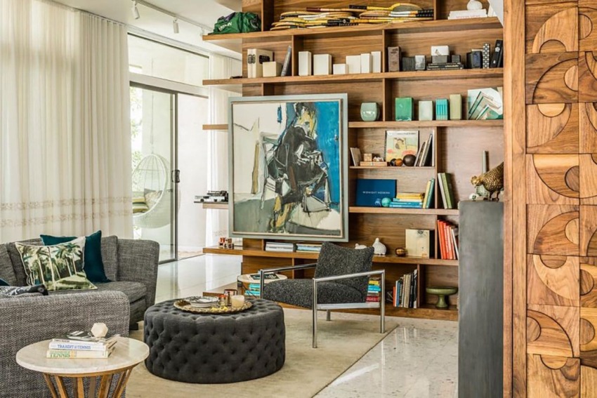 Modern living room with wooden shelves, abstract art, gray furniture, round ottoman, and glass doors leading outside.