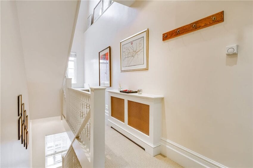 Bright hallway with white staircase, framed art on walls, and coat rack; carpeted floor and natural light from windows.