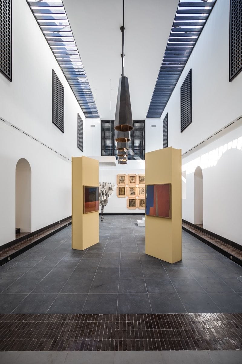 Modern art gallery interior with tall ceilings, hanging lights, skylights, and contemporary artworks on display.