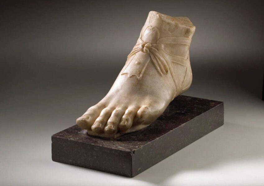 Marble sculpture of a realistic foot with a sandal, displayed on a dark rectangular base.
