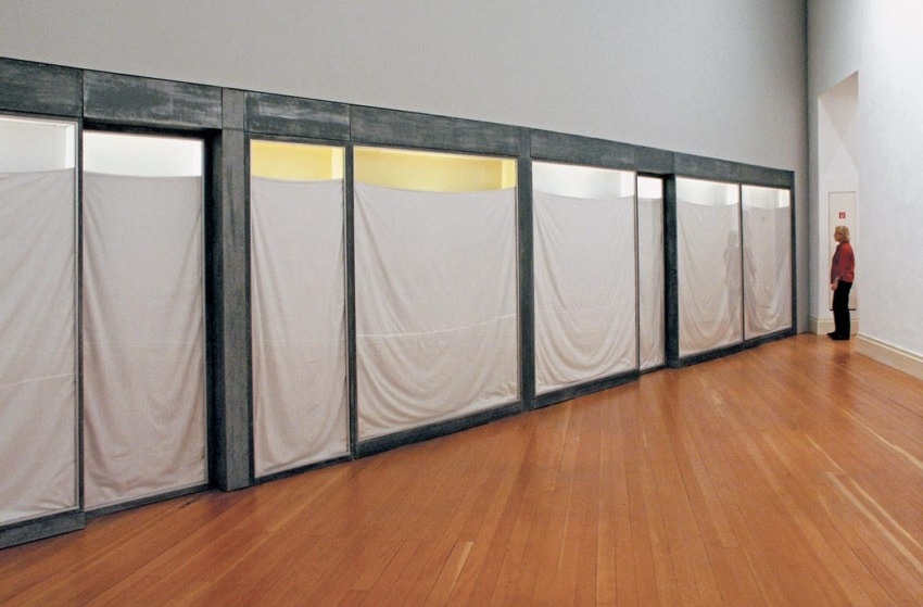 Art installation featuring a row of partitioned sections with white drapes and a person standing near a wall in a gallery space.