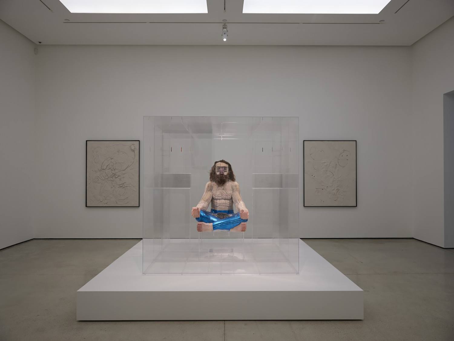 Art installation in a gallery featuring a person suspended in a transparent box, surrounded by abstract drawings.