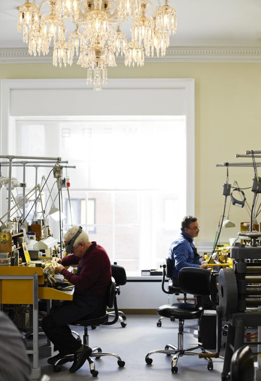 Workers crafting jewelry under a chandelier in a well-lit workshop with tools and equipment.