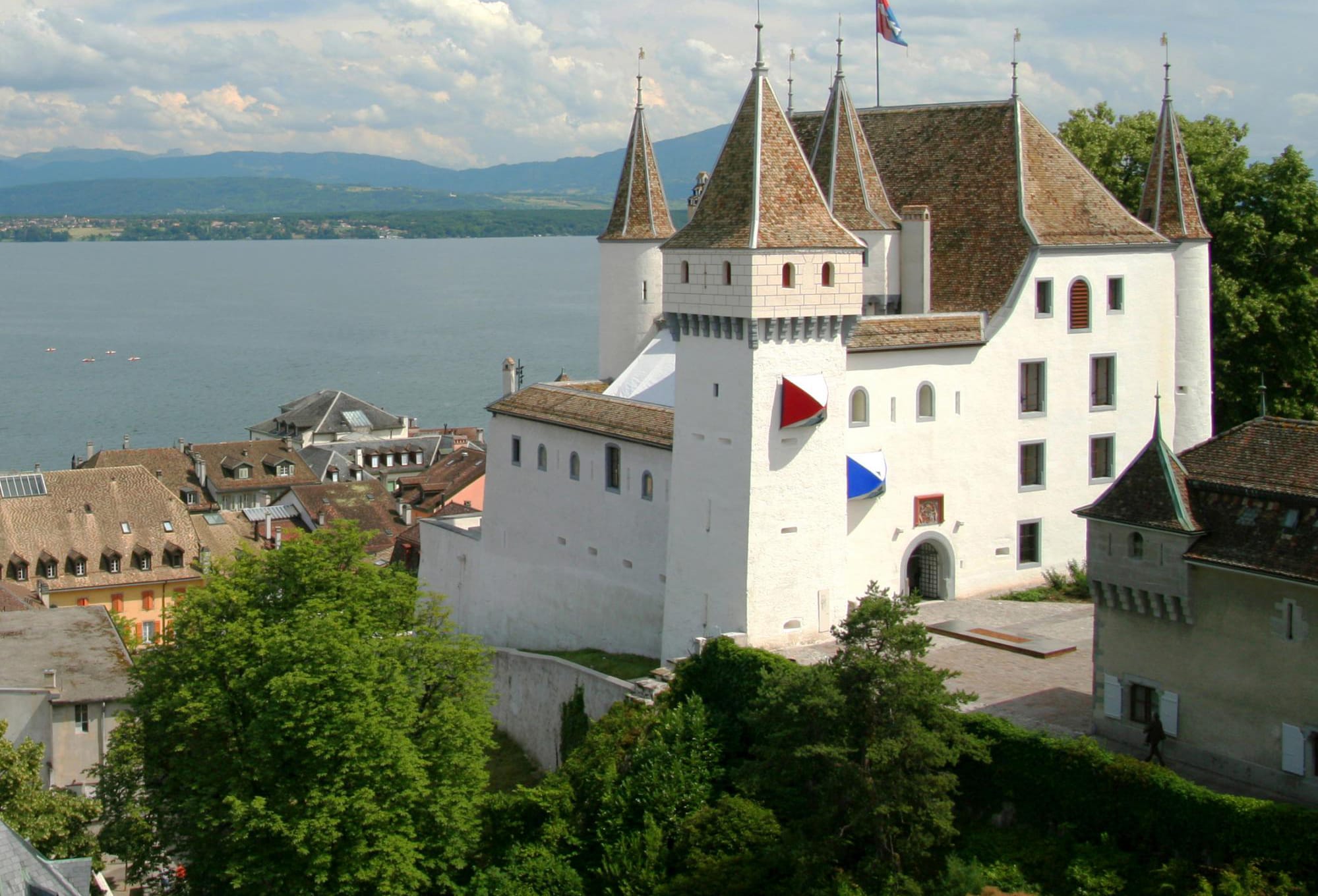 Castle with pointed towers near a lake, surrounded by trees and quaint village buildings under a partly cloudy sky.