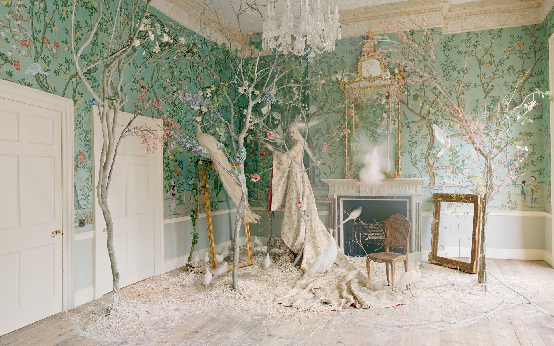 Surreal room with tree branches, birds, and floral wallpaper, featuring an empty chair and a draped figure near a fireplace.