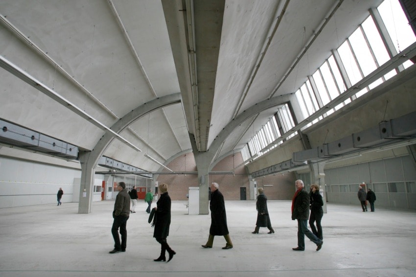 People walking in a large, empty industrial hall with high ceilings and arched beams.