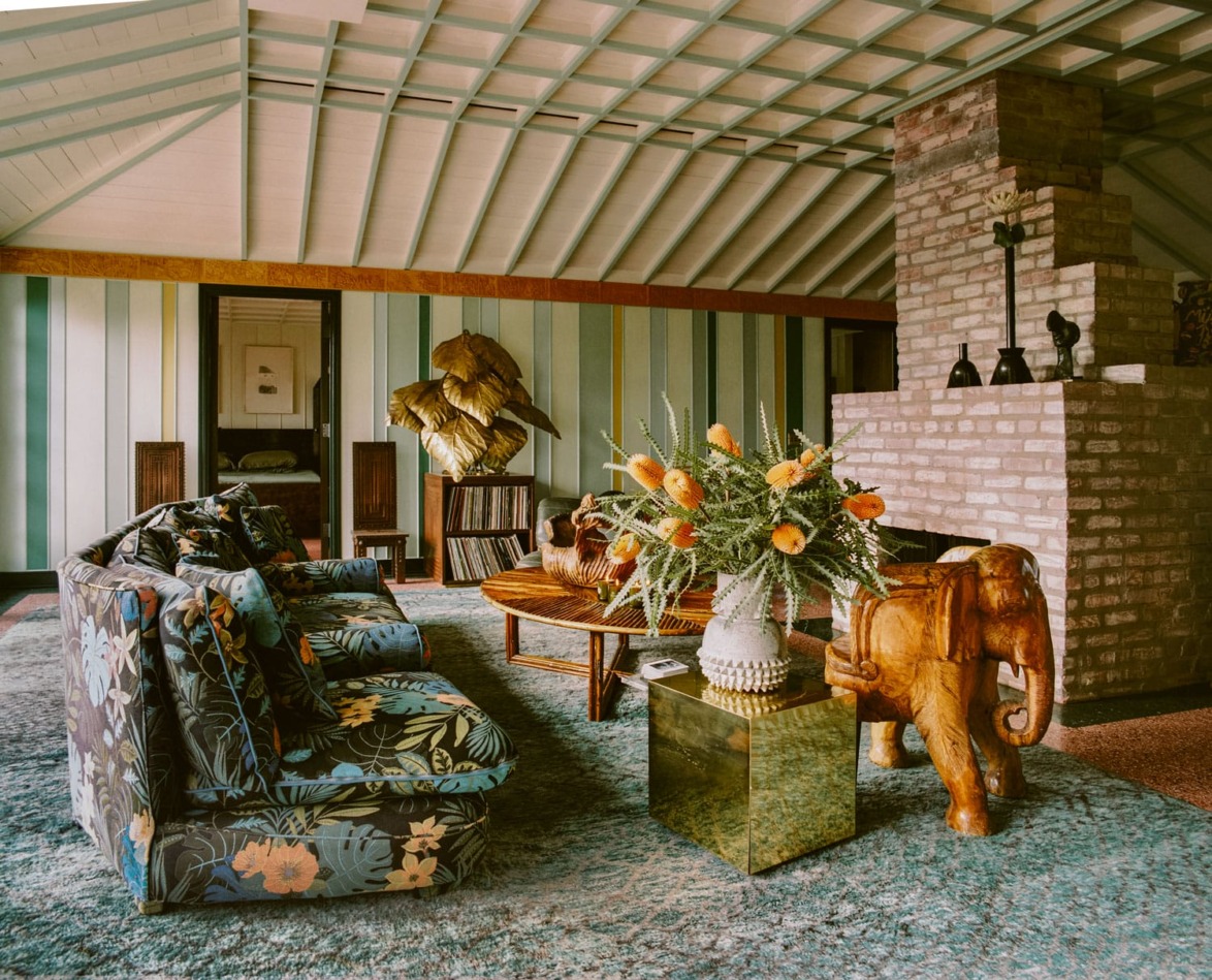 Retro-style living room with floral furniture, brick fireplace, decorative elephant sculpture, and vibrant floral arrangement.