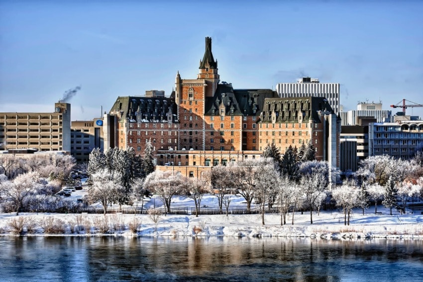 Grand historic hotel with winter landscape, tall central tower, surrounded by trees and snow, reflecting in a calm river.