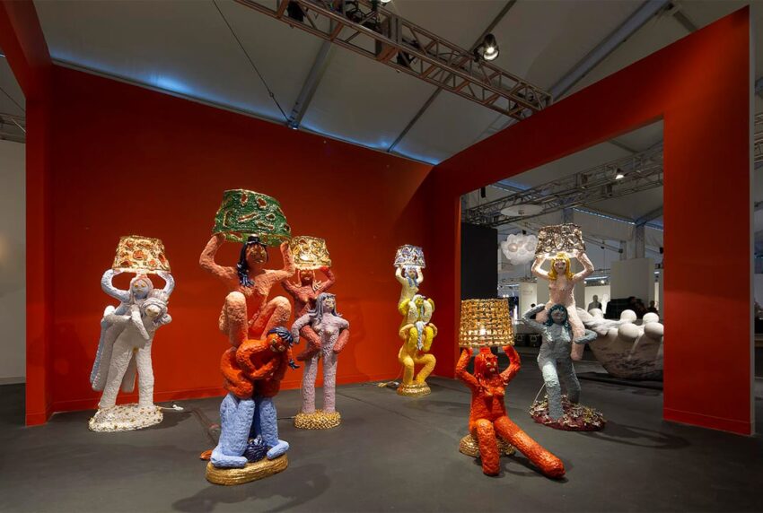Colorful abstract sculptures resembling people made from beads, each holding a unique basket on their head in an art exhibit.