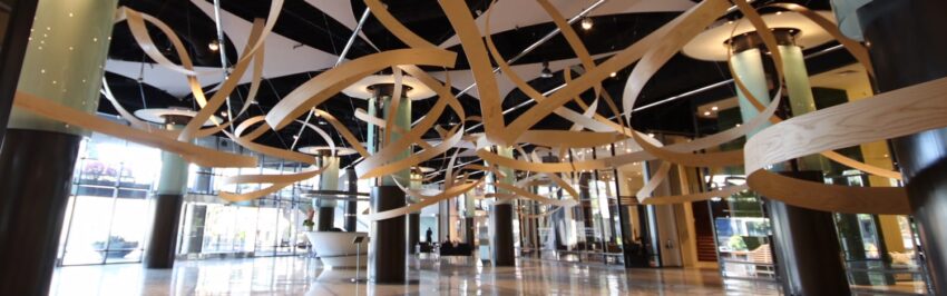 Modern lobby with artistic wooden ribbon ceiling design, glass walls, and polished floors illuminated by ceiling lights.