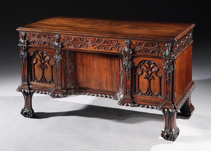 Antique wooden cabinet with intricate gothic carvings, featuring decorative arches and ornate leg design on a dark background.