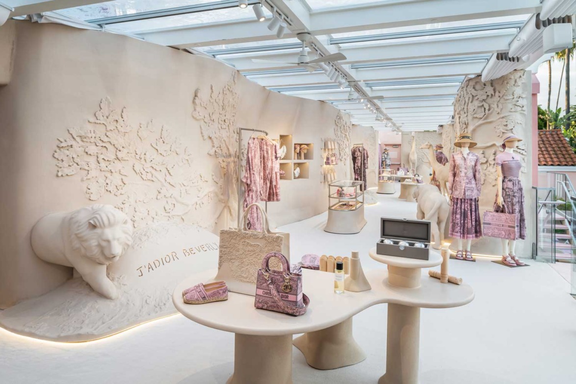 Elegant boutique interior with sculptures and displays of fashionable clothing, accessories, and decor under a glass ceiling.