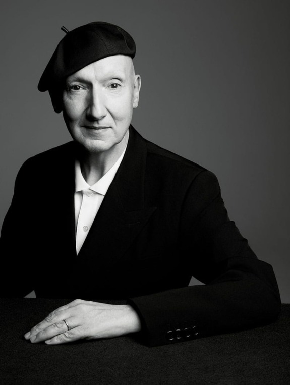 Person in black and white photo wearing a beret and suit, sitting at a table with hands folded, looking forward.