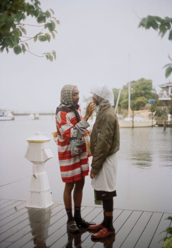 Two people standing on a dock near the water, wearing layered clothing and headscarves, surrounded by trees and boats.