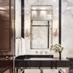 In a marble-lined powder room, custom sconces by Jonathan Browning from the Bright Group illuminate the custom-made vanity with Lefroy Brooks sink fixtures.