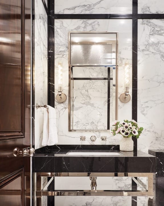 In a marble-lined powder room, custom sconces by Jonathan Browning from the Bright Group illuminate the custom-made vanity with Lefroy Brooks sink fixtures.