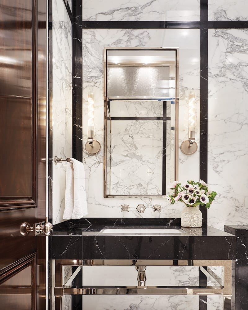 In a marble-lined powder room, custom sconces by Jonathan Browning from the Bright Group illuminate the custom-made vanity with Lefroy Brooks sink fixtures.
