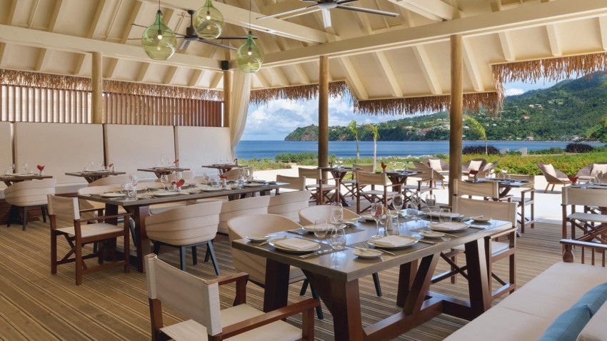 Open-air restaurant with ocean view, wooden tables, and white chairs under a thatched ceiling, set for dining.