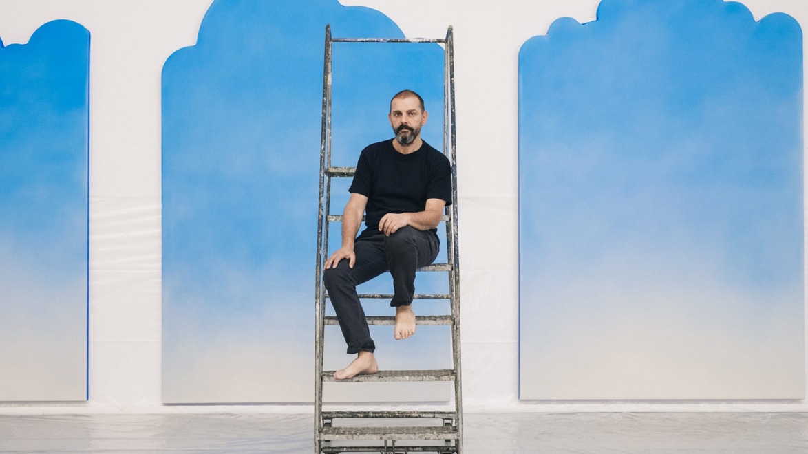 Man sitting on a metal ladder with blue cloud-like shapes in the background in a bright indoor setting.
