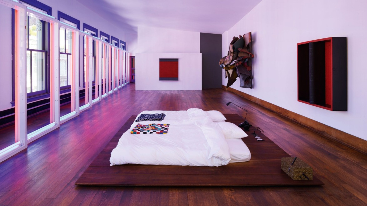 Modern art gallery with minimalist bed display, wooden floors, large windows, and colorful abstract wall art.