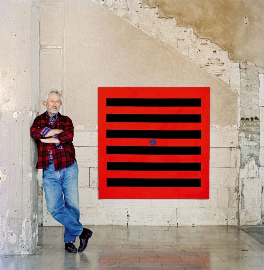 Man in plaid shirt leans against wall beside large square artwork with red and black stripes.