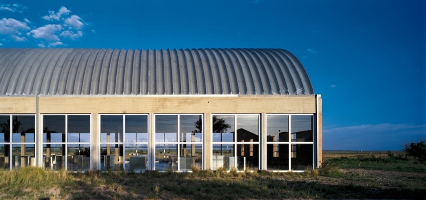 Modern building with curved metal roof and large glass windows reflecting the clear blue sky and grassy landscape.