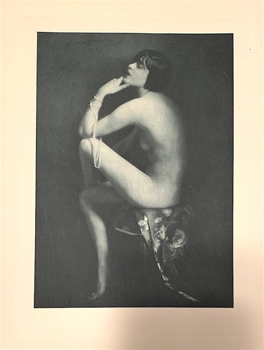 Black and white image of a seated woman in profile, draped in pearls, appearing contemplative against a dark backdrop.