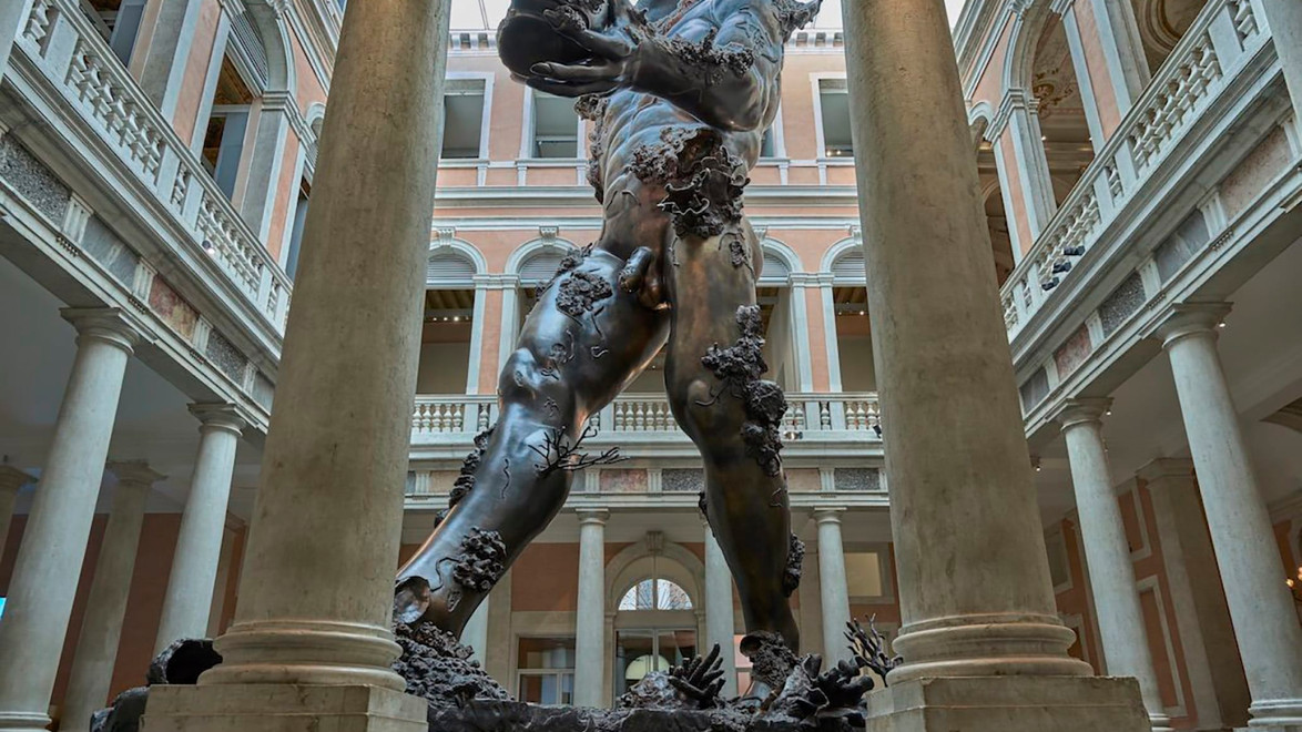 Giant bronze statue of a muscular figure with coral formations, displayed in an ornate architectural setting with columns.
