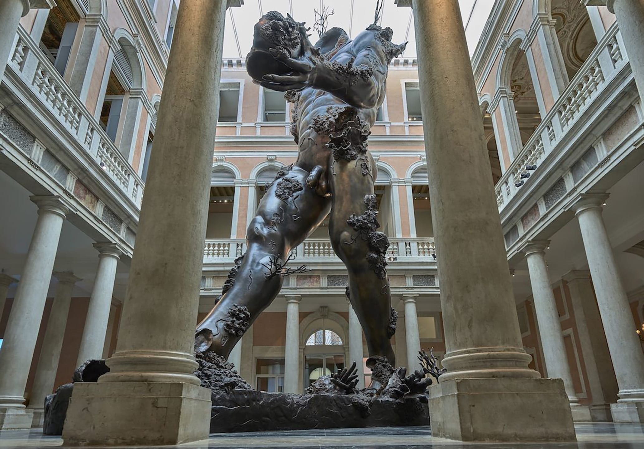 Giant bronze statue of a muscular figure with coral formations, displayed in an ornate architectural setting with columns.