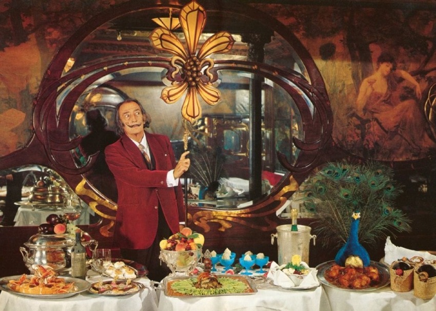Man in a red suit poses with an ornate interior backdrop and a lavish table filled with a variety of foods and decorations.