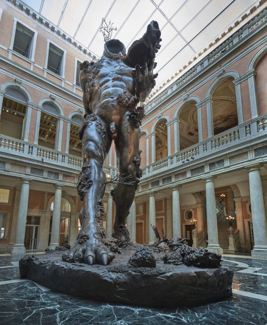 Massive headless bronze sculpture of a muscular figure in an ornate museum atrium with marble columns and arched windows.